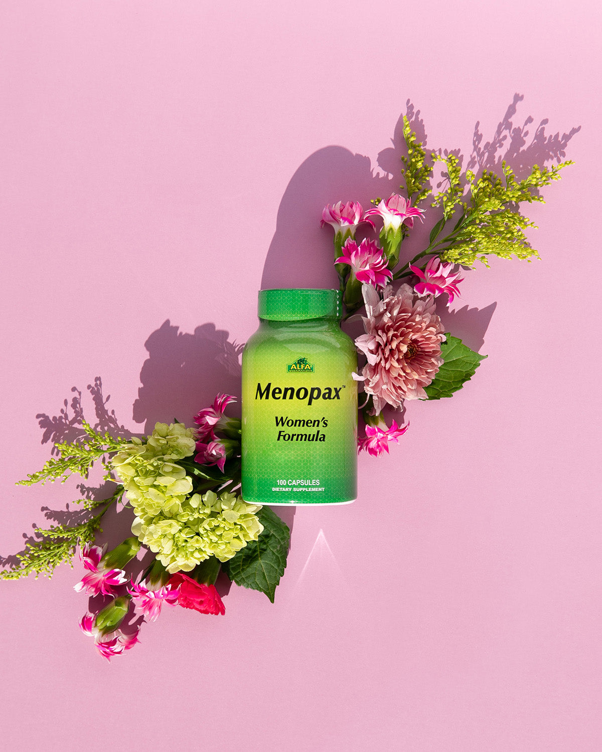 A green bottle of Alfa Vitamins Menopax Womens Formula - 100 capsules is stylishly displayed among pink, white, and green flowers and leaves on a pink background, highlighting its commitment to menopause support and hormonal balance.