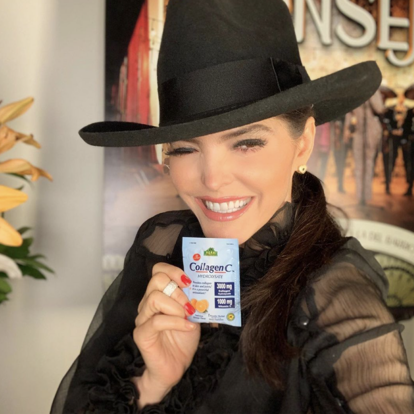 A person in a black hat and blouse smiles, holding Alfa Vitamins CollagenC Hydrolysate powder with Vitamin C - 30 Pack, recognized for boosting skin elasticity. Flowers and a blurred poster create a charming backdrop.