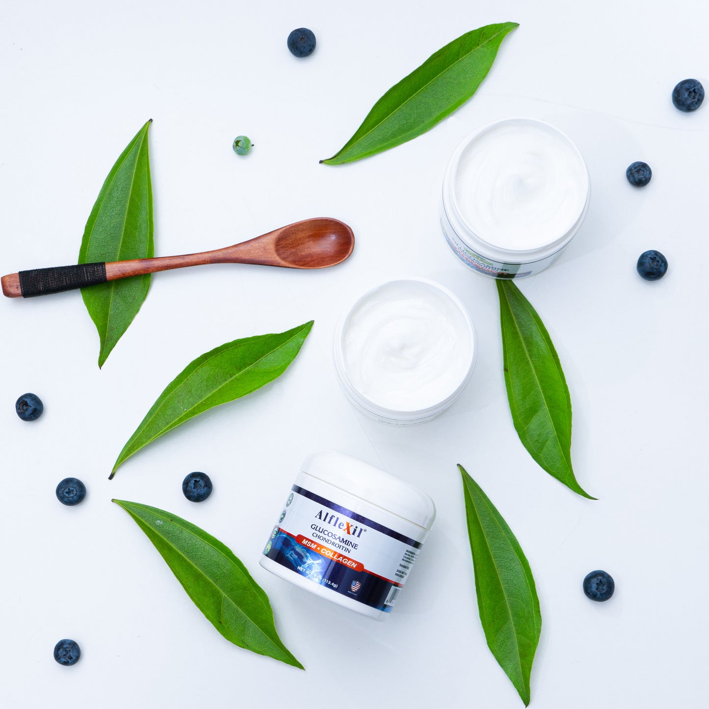 Three jars of ALFLEXIL® Glucosamine + Chondroitin Cream by Alfa Vitamins for joint relief are placed on a white background with green leaves and blueberries surrounding them, accompanied by a wooden spoon.