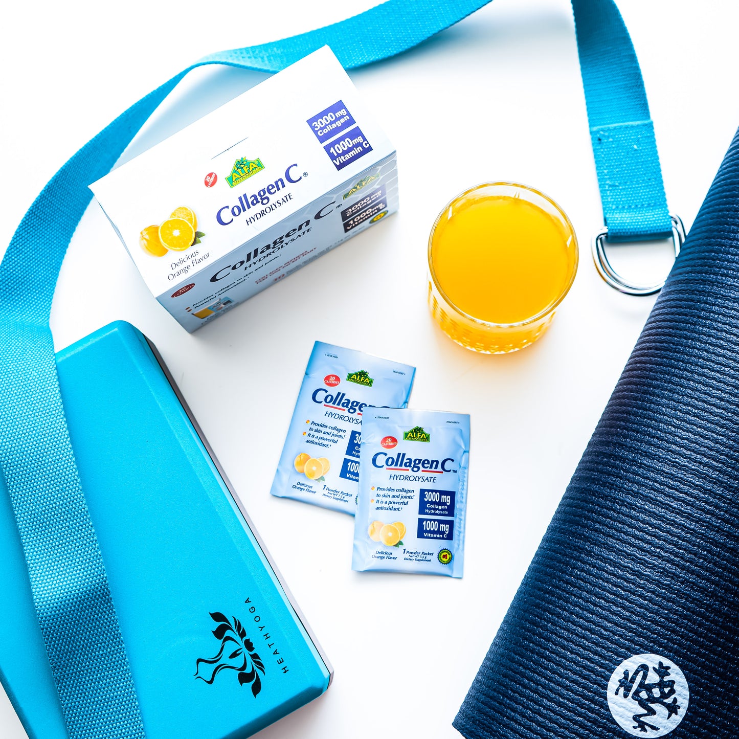 A flat lay displays Alfa Vitamins CollagenC Hydrolysate powder with Vitamin C - 30 Pack, a blue water bottle, orange juice, and a rolled-up yoga mat, all neatly arranged on a clean white surface.