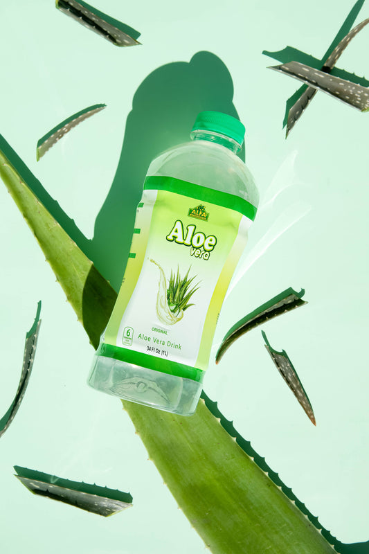 An Aloe Vera Drink-Original by Alfa Vitamins rests on a green surface amid aloe leaves. The bottle, with its green cap and Aloe Vera label featuring the plants image, creates a dynamic play of light and shadow while serving as a superfood elixir that supports the digestive system.