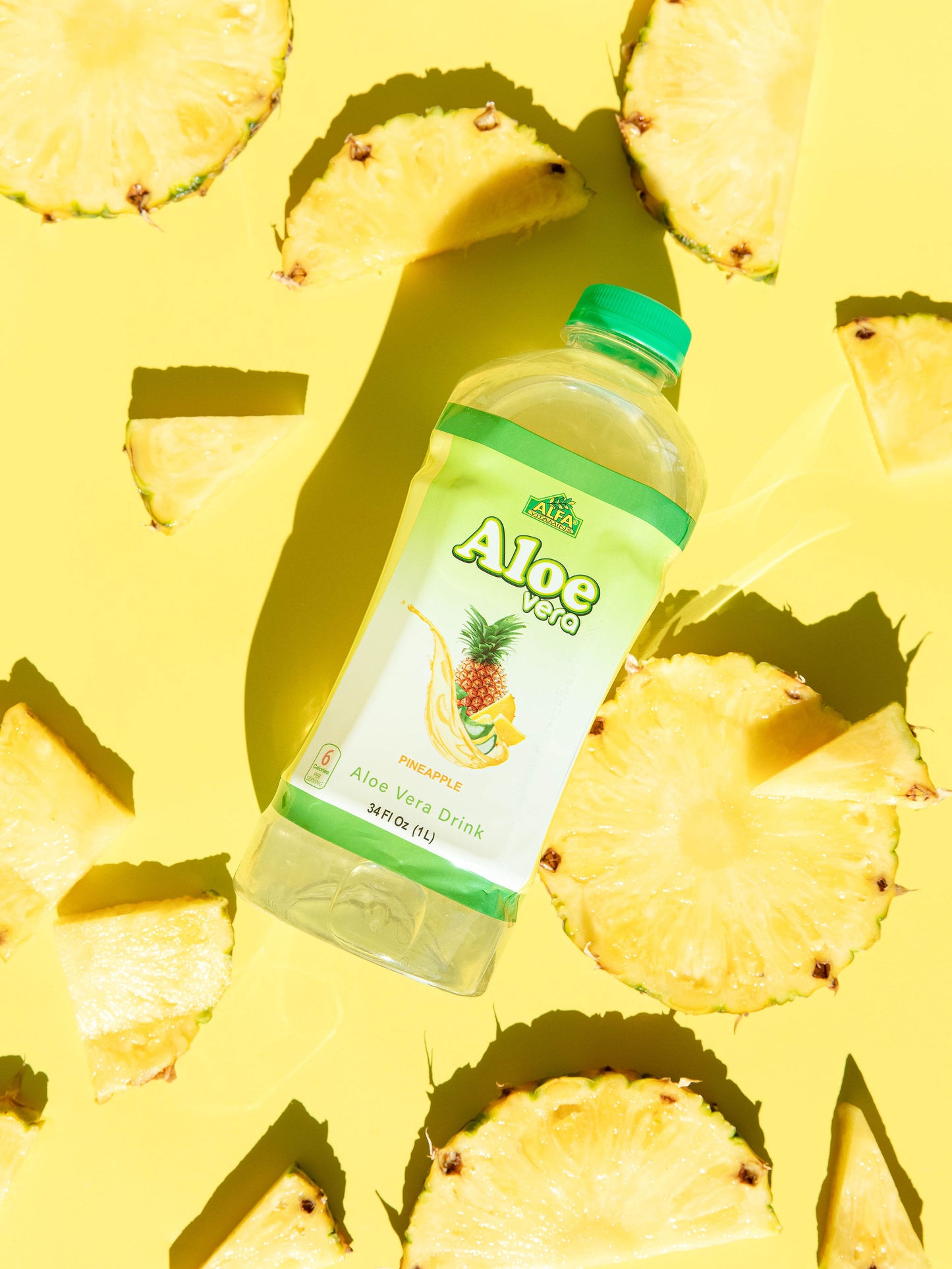 On a bright yellow background, an Alfa Vitamins Aloe Vera Drink-Pineapple bottle sits surrounded by sliced pineapple. The drink, known for digestive support, features a green cap and a label with a pineapple image, creating an ideal superfood elixir scene.