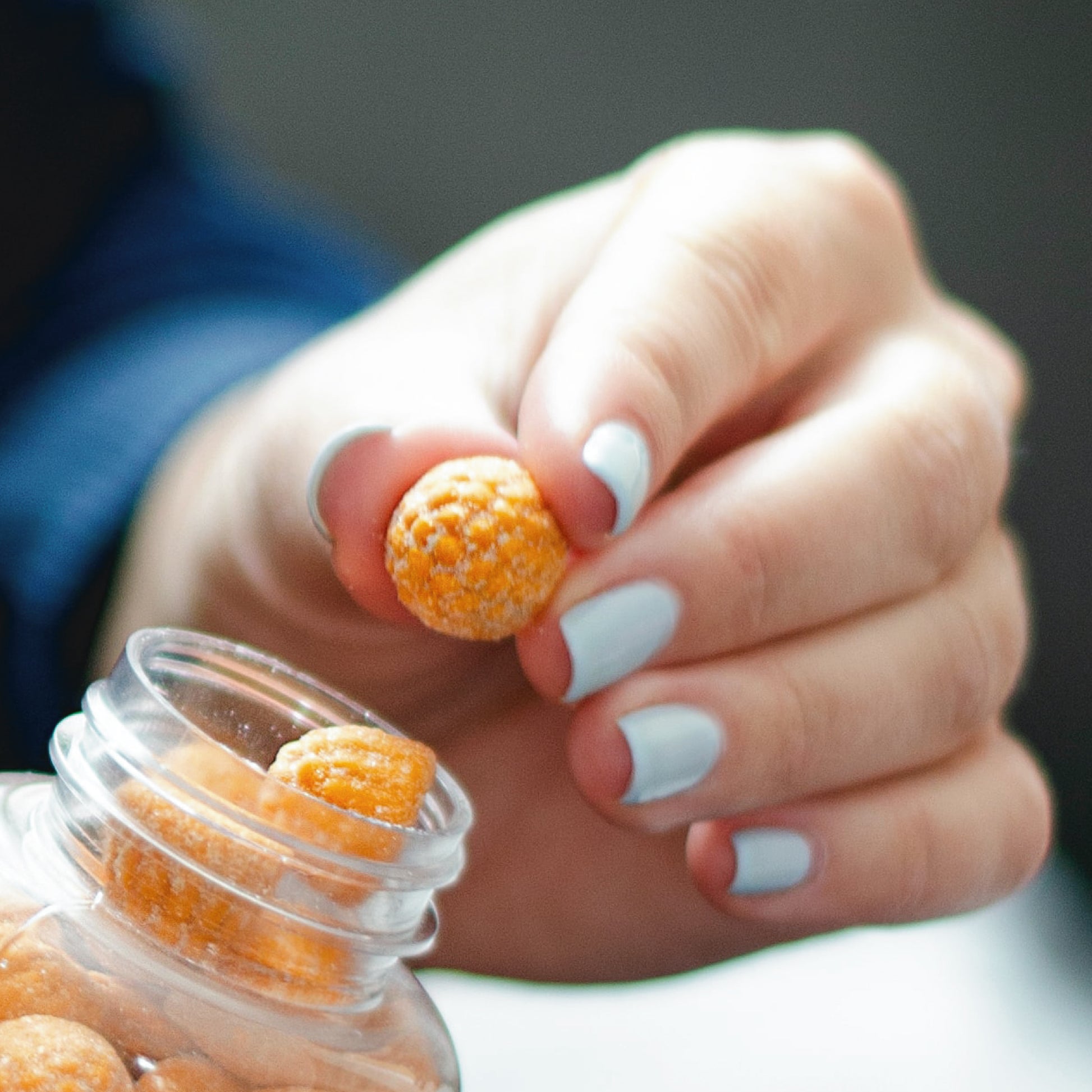 A hand with light blue nails holds an orange Turmeric Ginger Gummy from Alfa Vitamins Store, taken from a transparent jar labeled 60 Gummies, against a blurred background.