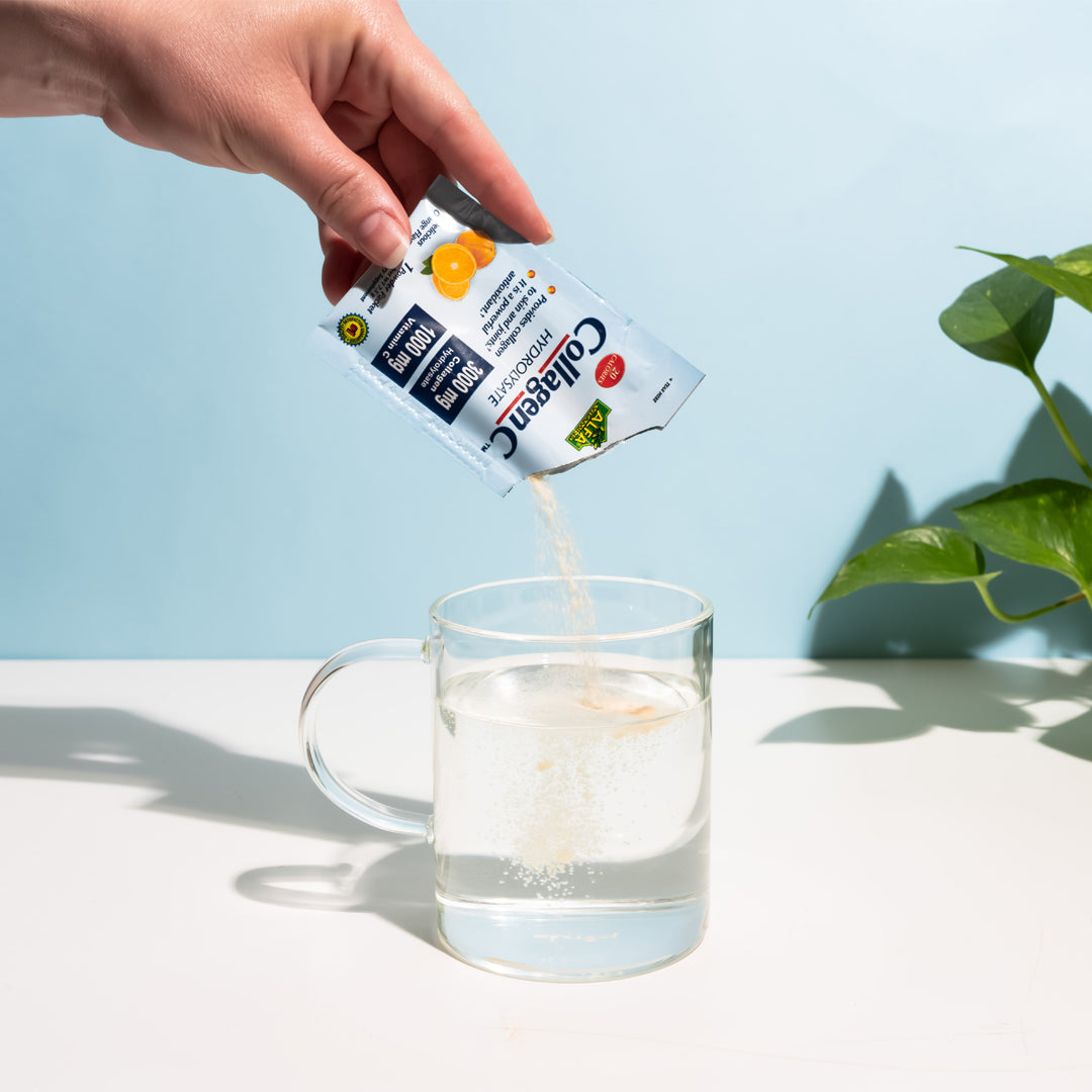 A hand pours a packet of Alfa Vitamins CollagenC Hydrolysate powder with Vitamin C into a clear mug of water. The packaging highlights protein, energy, and immune boost. Promoting skin elasticity, it sits on a light blue surface with a green plant in the background.