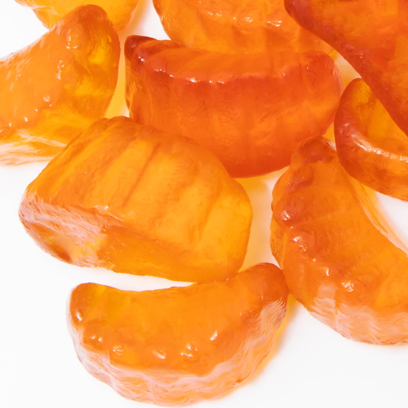A close-up of Alfa Vitamins Vitamin C Gummies for Adults (60 gummies) reveals translucent orange candies shaped like orange slices with a shiny, ridged surface. These gummies resemble real fruit segments and are scattered randomly against a plain white background.