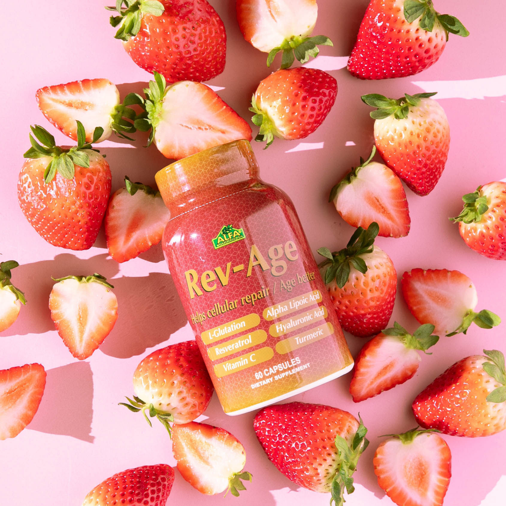 A bottle of Alfa Vitamins REV-AGE Cellular Repair Formula, containing 60 capsules, sits on a pink surface surrounded by fresh strawberries. It features antioxidant ingredients like Glutathione, Resveratrol, and Turmeric for cellular regeneration and anti-aging benefits.