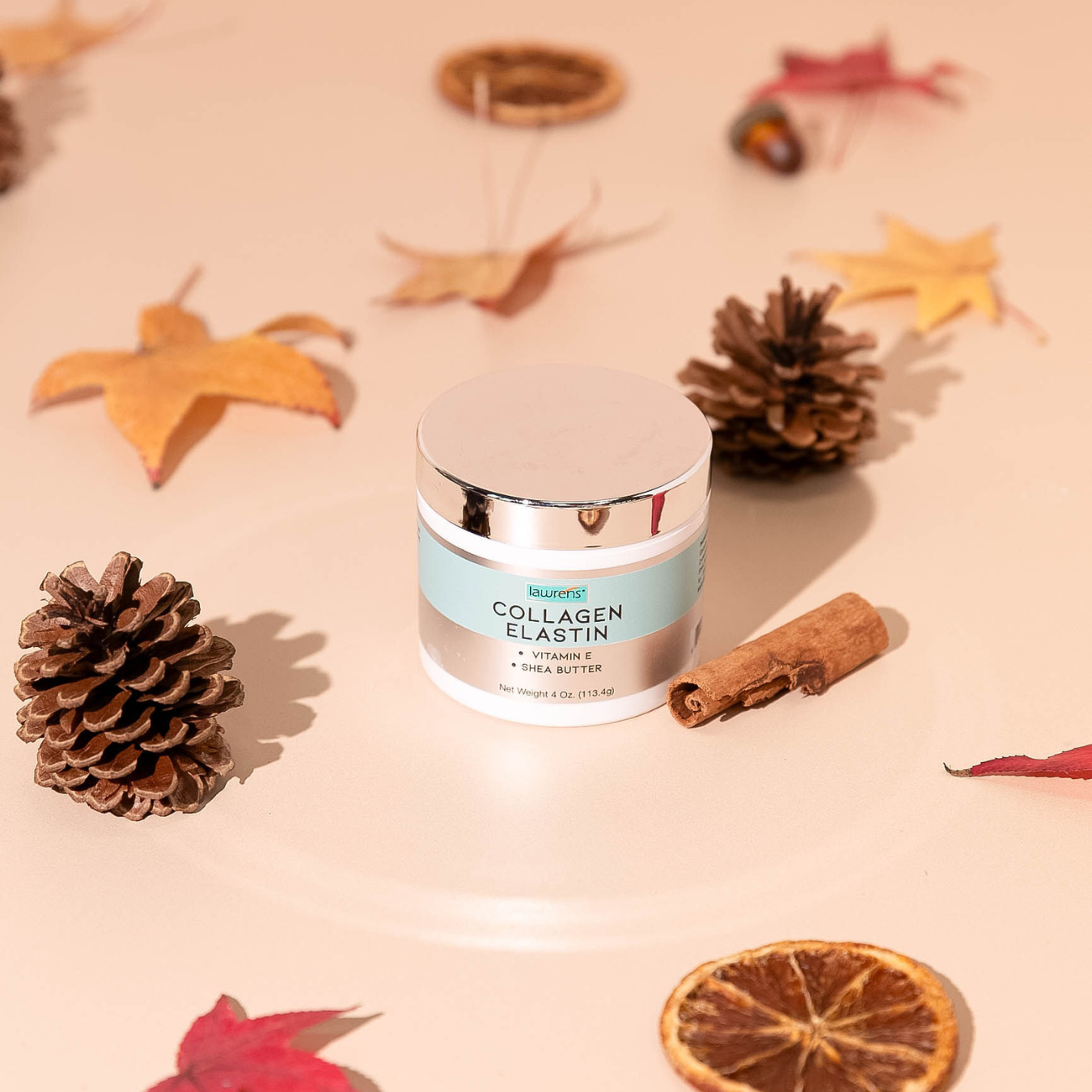 The 4 oz jar of Alfa Vitamins Collagen Elastin Cream rests on a beige surface, promising youthful skin. Surrounded by autumn leaves, pinecones, a cinnamon stick, and dried citrus slices, it creates a warm seasonal vibe reflecting its skin elasticity benefits.