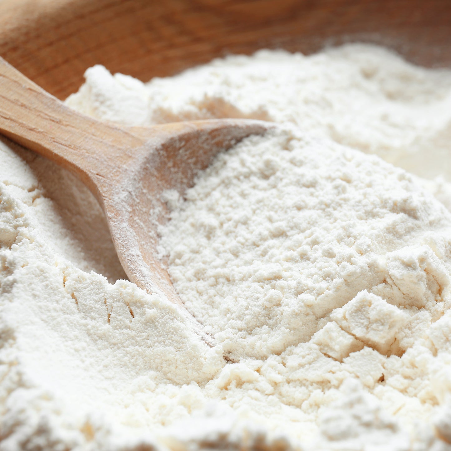 A wooden spoon sits in a pile of flour, highlighting its powdery texture. The rustic background subtly suggests using Alfa Vitamins Maximum Whey Protein Powder for added richness.