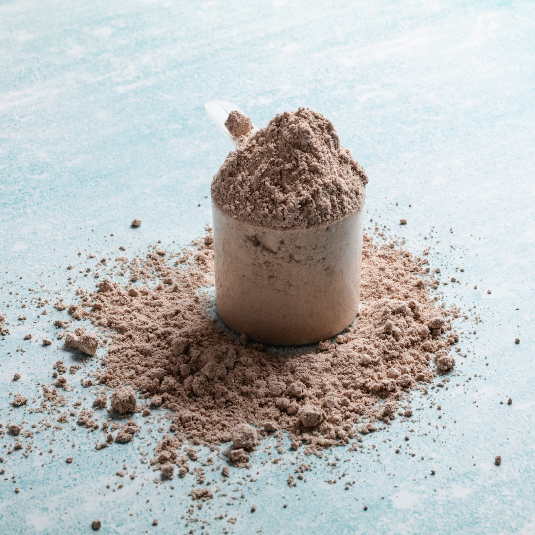 A clear scoop of Alfa Vitamins Maximum Whey Protein Powder (Chocolate, 2 lbs) sits on a light blue surface with scattered powder around it, the handle visible. The textured powder suggests essential amino acids vital for muscle growth.