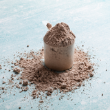 A clear scoop of Alfa Vitamins Maximum Whey Protein Powder (Chocolate, 2 lbs) sits on a light blue surface with scattered powder around it, the handle visible. The textured powder suggests essential amino acids vital for muscle growth.