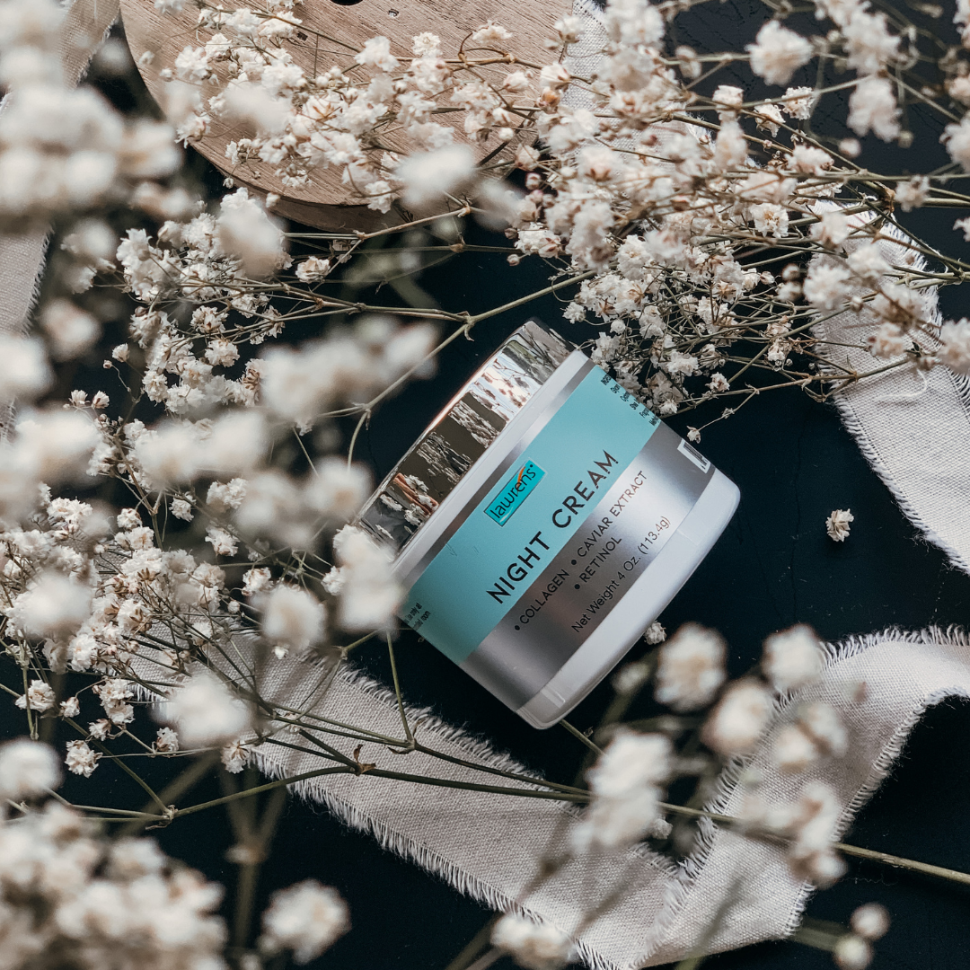 A jar of Alfa Vitamins Night Cream with Collagen, Caviar Extract & Retinol - 4 oz rests on a black surface, surrounded by delicate white flowers and zigzagging fabric strips, creating a serene and elegant arrangement.