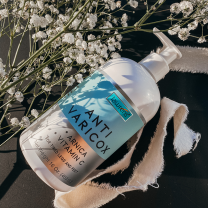 A 16 oz bottle of Lawrens Cosmetics Anti-Varicox Cream by Alfa Vitamins Store rests on a black surface, surrounded by white flowers and beige fabric. Designed for tired legs and feet.