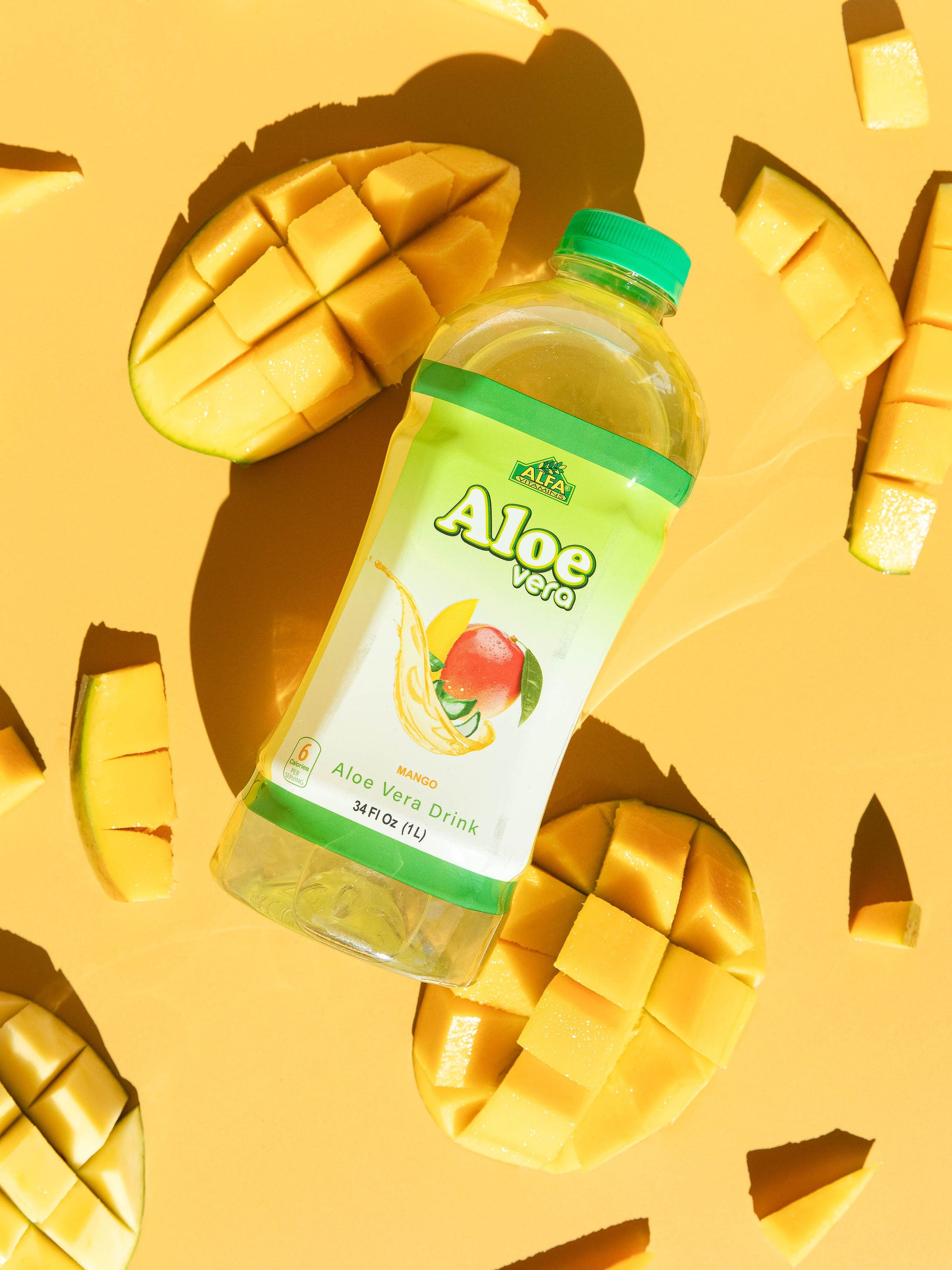 The Aloe Vera Drink-Mango by Alfa Vitamins, a superfood elixir for digestive support, is set against a bright yellow background with sliced mango pieces. The bottle showcases a vibrant mango graphic alongside aloe vera leaves.