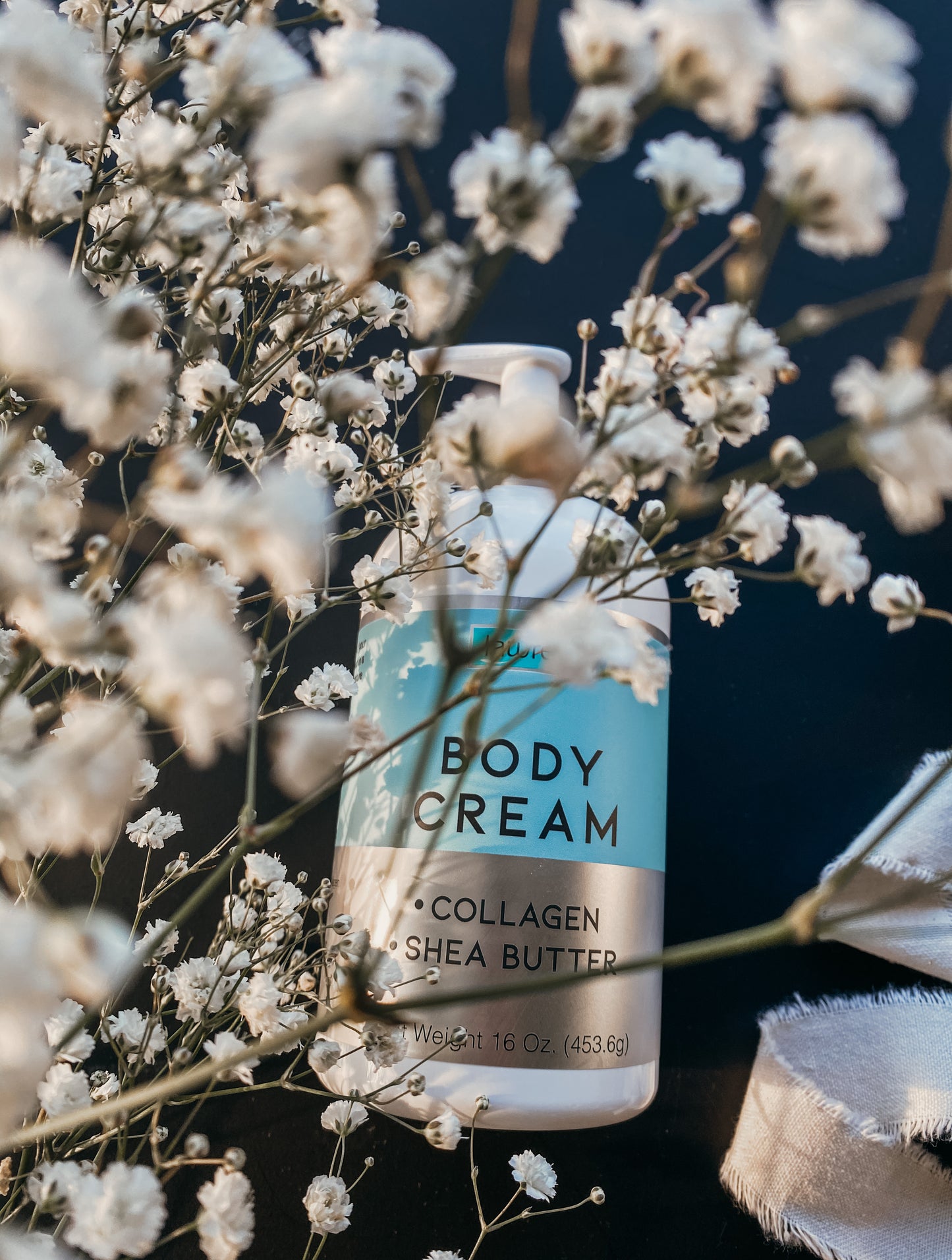 A 16 oz bottle of Body Cream Collagen with Shea Butter by Lawrens Cosmetics from Alfa Vitamins rests elegantly among delicate white babys breath flowers and a piece of fabric against a dark background, offering hydration and anti-aging benefits with its nourishing formula.