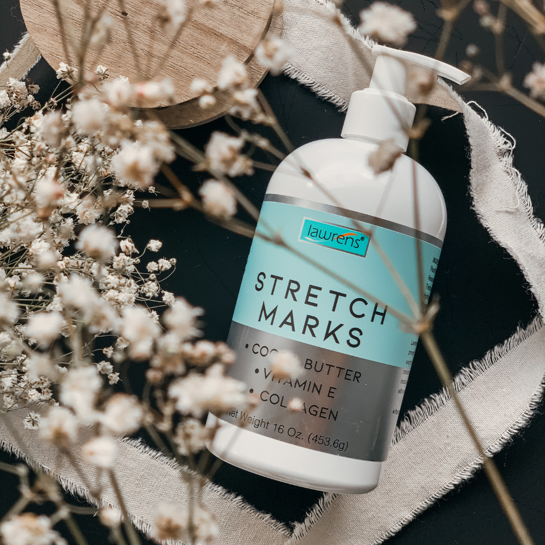 A 16 oz bottle of Alfa Vitamins Stretch Marks Cream with Cocoa Butter, featuring a pump and natural ingredients like cocoa butter, vitamin E, and collagen, rests on a black surface surrounded by delicate white flowers and a textured circular cloth.