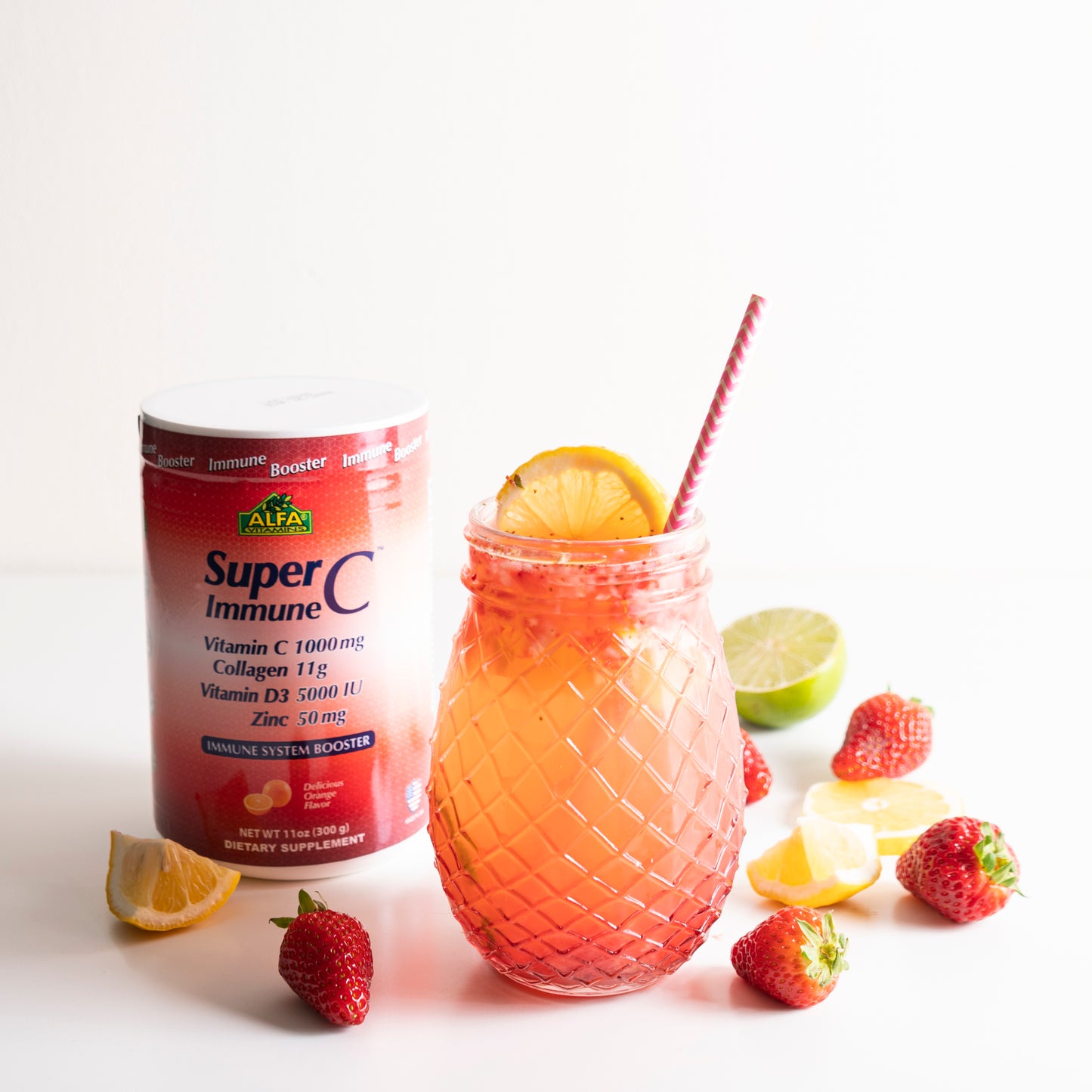 A mason jar with a pink straw and lemon garnish, surrounded by strawberries and lemon wedges, contains a refreshing beverage. Nearby is an Alfa Vitamins container labeled SuperC Immune powder formula - Orange Flavor 11oz, highlighting its immune-boosting role. A lime sits in the background.