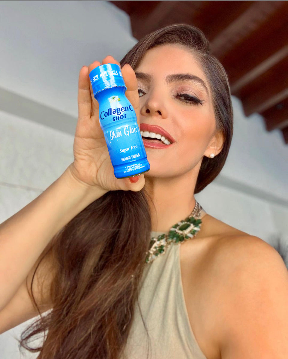 A woman with long hair holds a blue CollagenC Shot - Skin Glow bottle from Alfa Vitamins Store, promising skin elasticity. She smiles in a sleeveless top with a beaded neckline, set against white walls and a wooden ceiling.